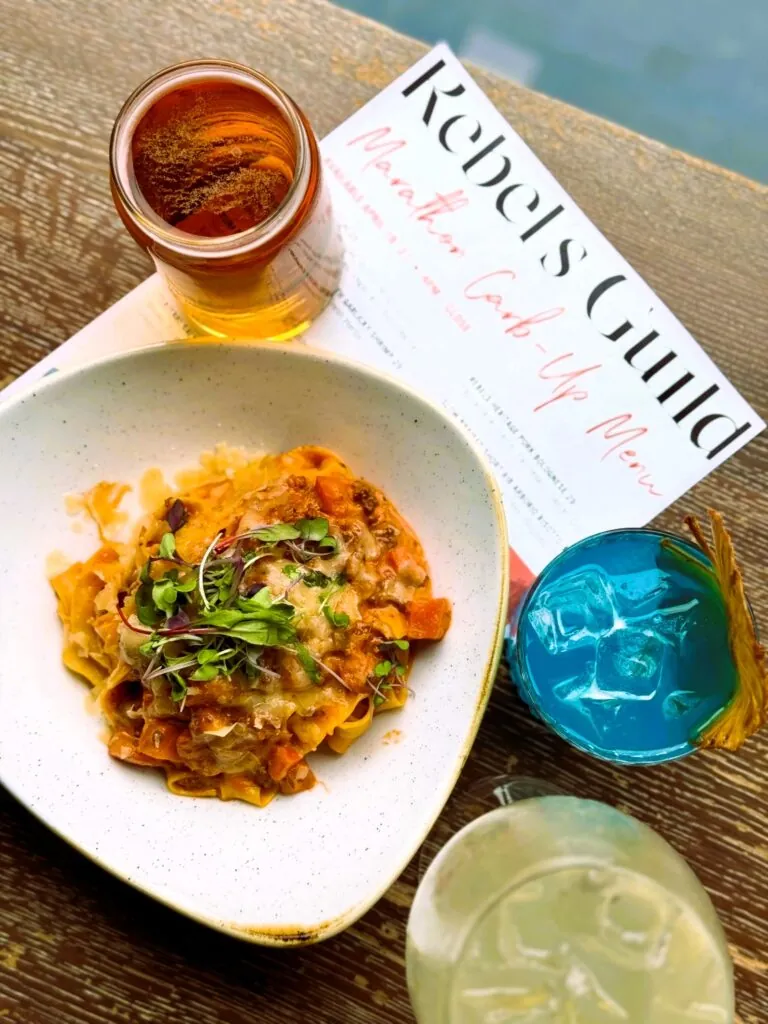 A plate of pasta with herbs, three colorful drinks, and a Rebel’s Guild pop-up menu on a wooden table.