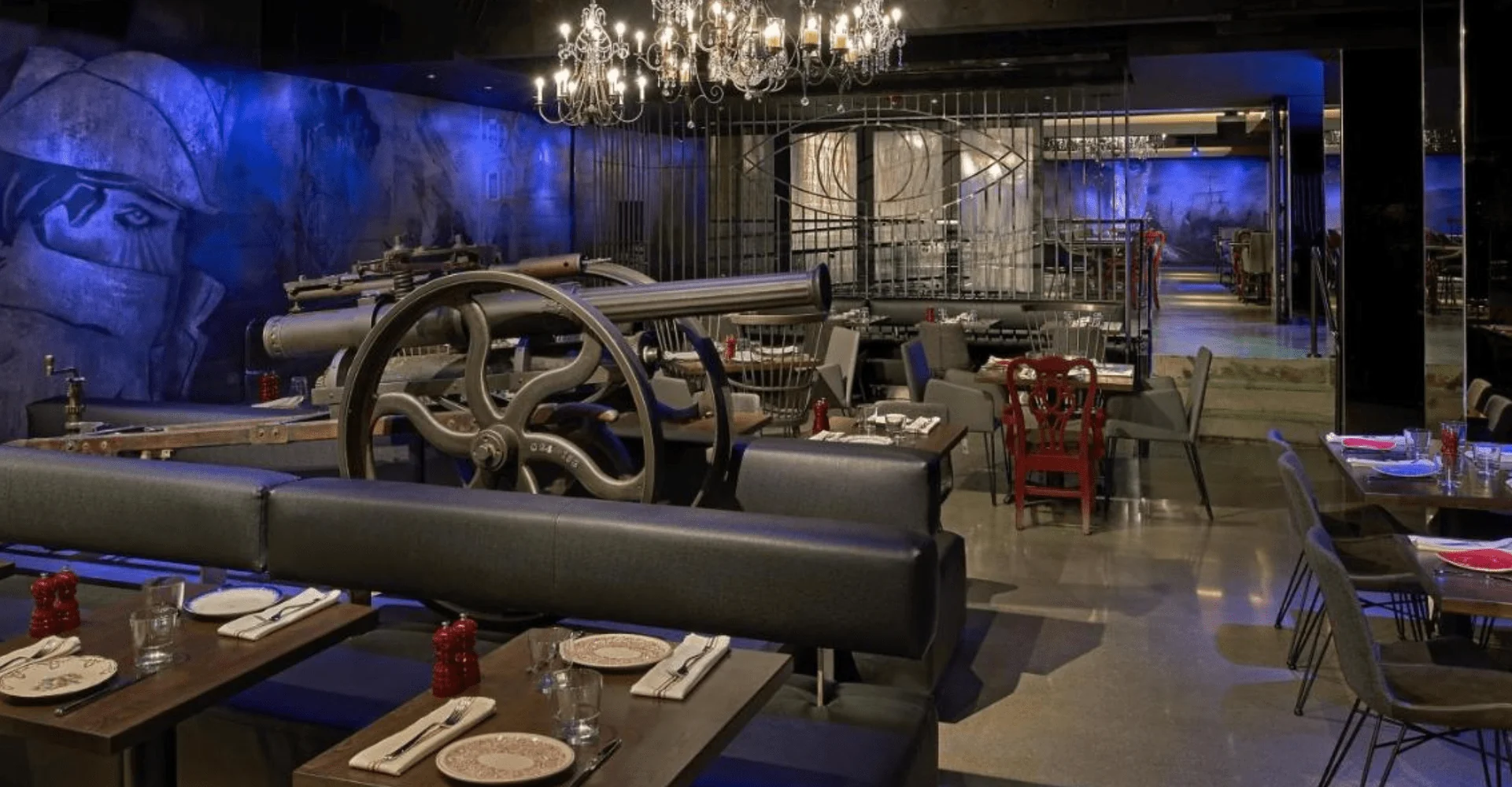 A dimly lit restaurant with industrial decor, like those in luxury hotels in Boston, features a large cannon, blue lighting, and striking murals.